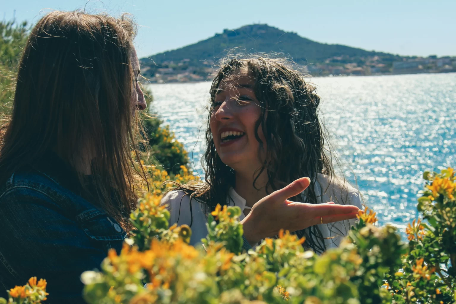 Two women standing and laughing in a field of flowers next to the ocean. 