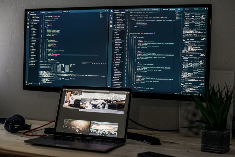 : An open laptop with a large monitor behind it, screen filled with code