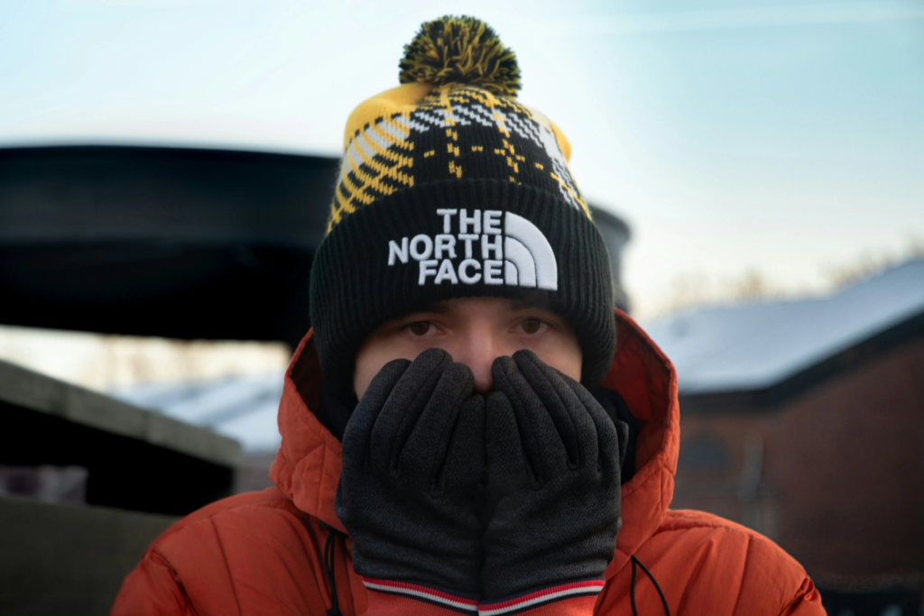 A man in a The North Face hat, standing in full winter gear with gloved hands over his face. 