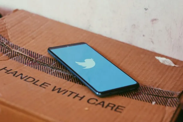 A smartphone displaying the Twitter logo is placed on a cardboard box labeled "HANDLE WITH CARE."