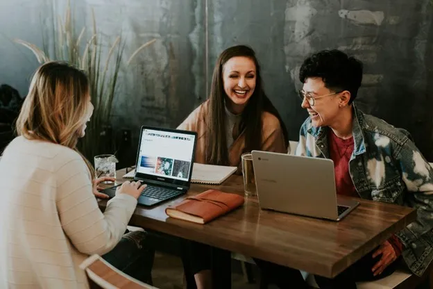 Three people are sitting around a table, working on their laptops and laughing together.