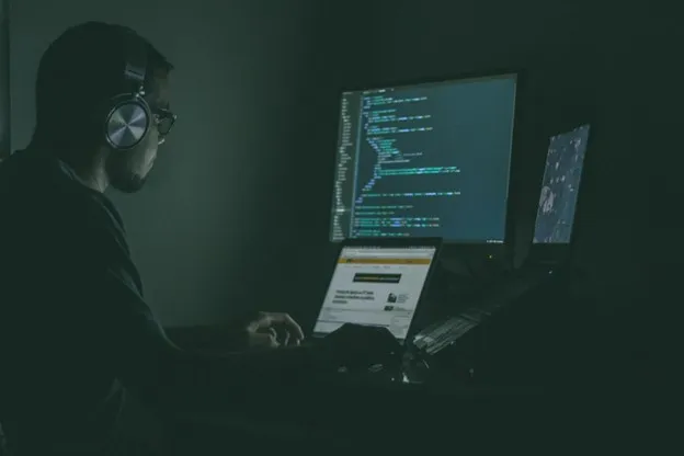 A hooded figure works on a laptop in a dark room, illuminated by a screen full of code.