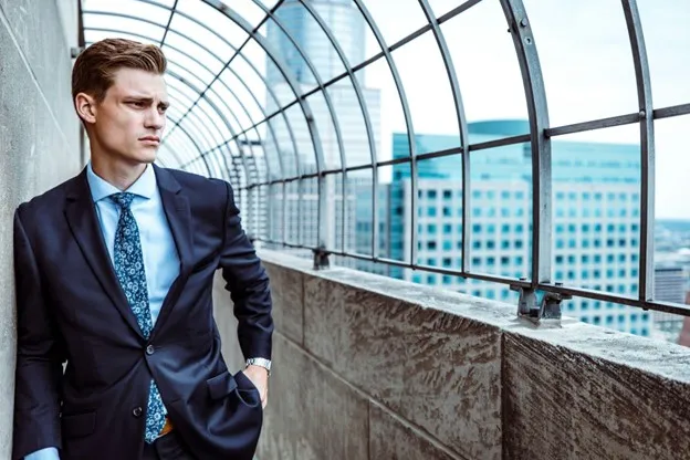 A man in a suit stands pensively on a rooftop with a cityscape in the background.