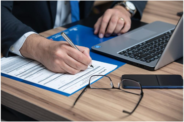 A person filling out paperwork at a desk, with a laptop, glasses, and smartphone nearby, indicating administrative or legal work.