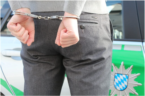 A person standing with hands cuffed behind their back, in front of a police vehicle.