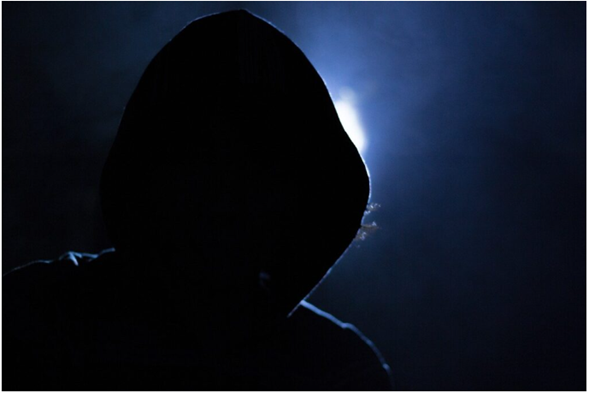 A mysterious person wearing a hoodie, silhouetted against a dim light in a dark environment, creating an eerie or secretive atmosphere.