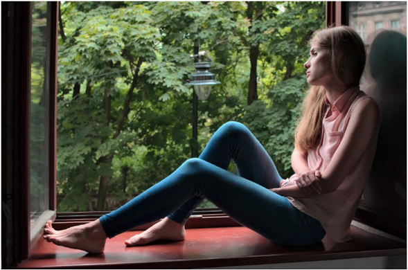 A young woman sitting by a window, gazing outside thoughtfully into a green landscape, appearing calm and reflective.